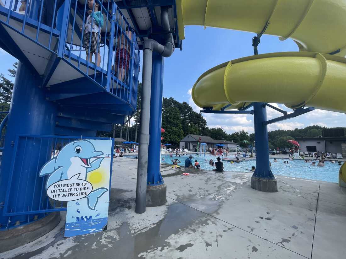 Muitas pessoas estão se refrescando na piscina azul-clara do Wills Park em Alpharetta, Geórgia.  A piscina tem um toboágua amarelo sinuoso e uma placa perto do toboágua dizendo que você deve ter pelo menos 48 centímetros de altura para andar nele.