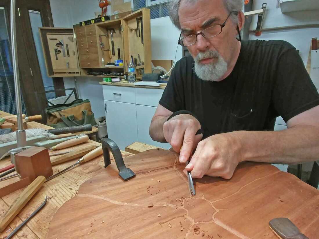 Robert Lang está sentado em um estúdio de marcenaria com muitas ferramentas de marcenaria ao seu redor. Ele segura uma ferramenta e a usa para esculpir uma grande placa de madeira que está apoiada em uma superfície.