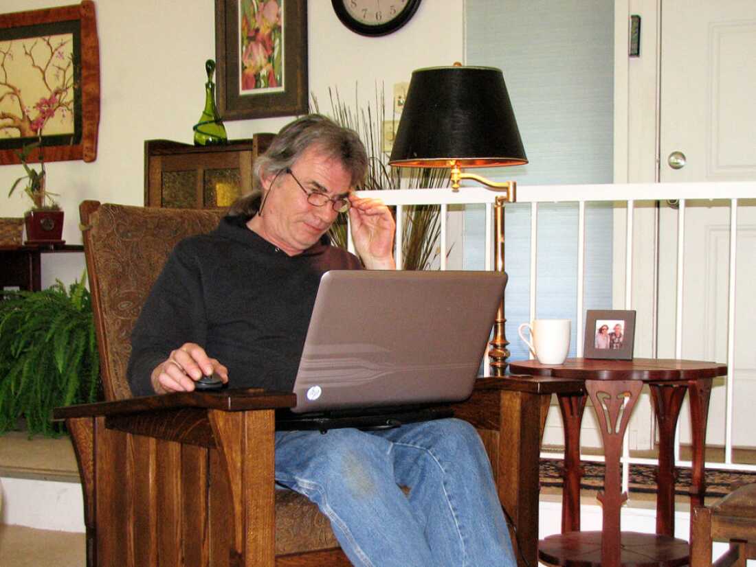 Robert Lang está sentado em uma poltrona com um laptop descansando em seu colo. Uma de suas mãos está em um mouse de computador que fica em um dos braços da poltrona.
