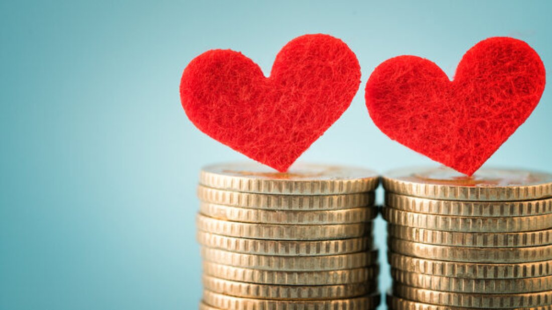 Dois corações de feltro vermelhos se equilibram sobre duas pilhas separadas de moedas de ouro contra um fundo azul claro, representando pessoas em um relacionamento romântico que funde finanças.